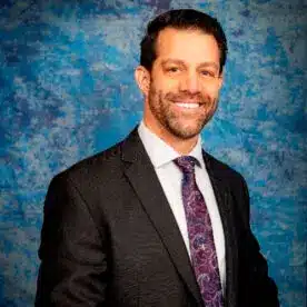 A man with short dark hair and a beard is smiling at the camera. He is wearing a dark blue suit, a checkered shirt, and a black tie. The background is a textured blue wall.